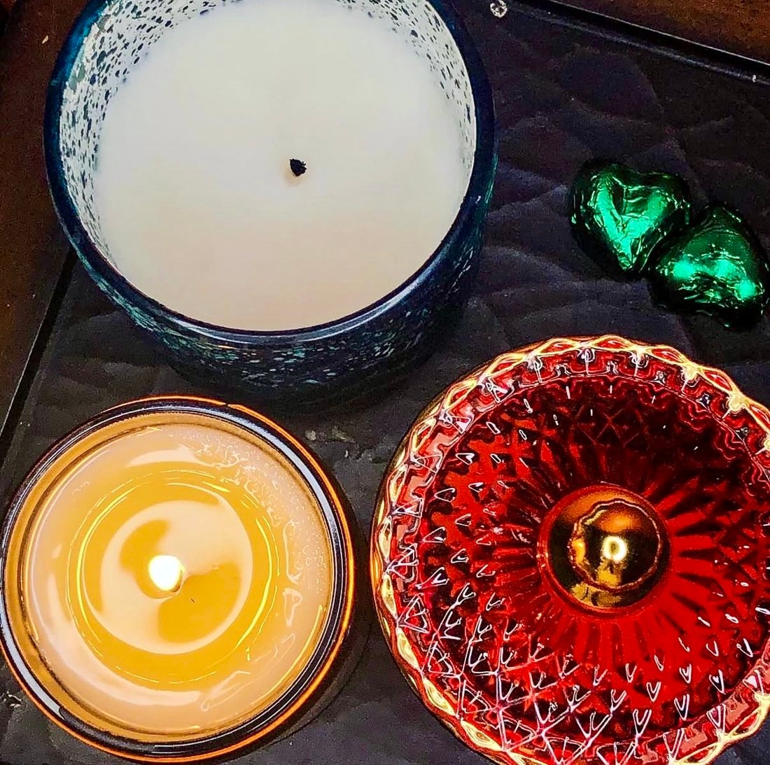 Two of Leah’s first candles seen from above — one in a speckled blue jar, one lit in an amber jar — beside a red glass dish and green foil-wrapped chocolates.