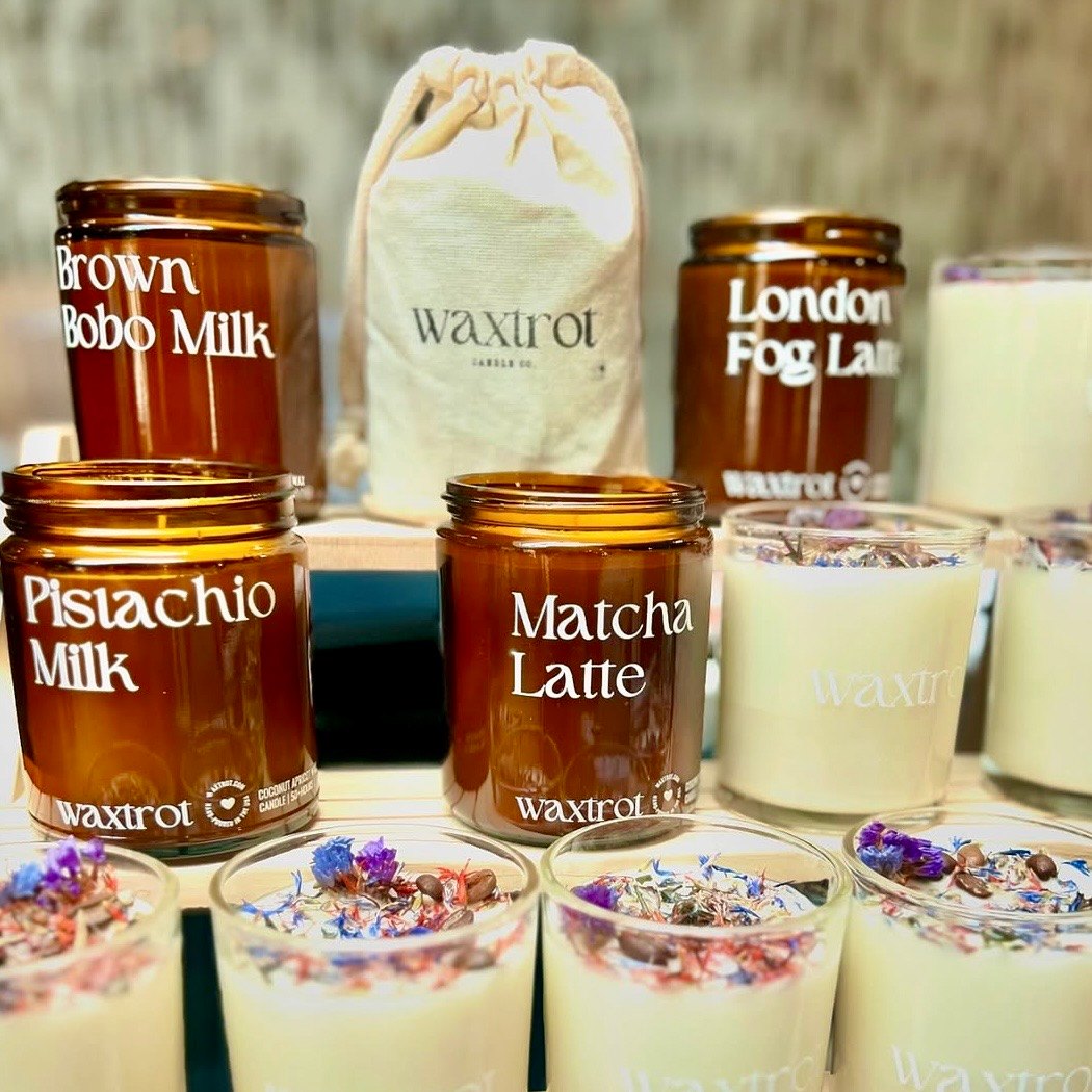 Candles on a market table: amber jars labelled Pistachio Milk, Matcha Latte and London Fog Latte, white jars topped with dried flowers, and a canvas gift pouch.