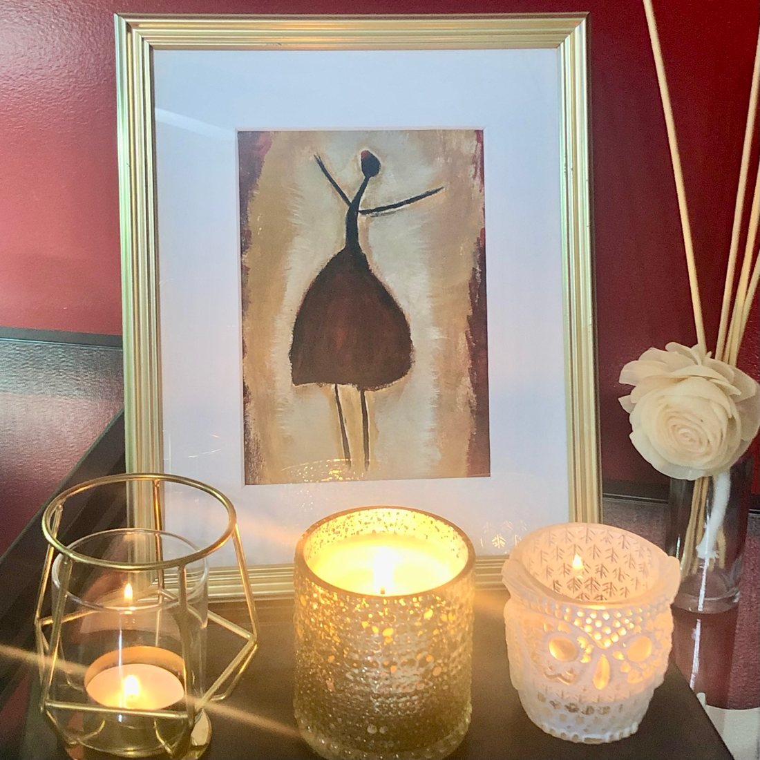Three lit candles in decorative holders in front of a framed painting.