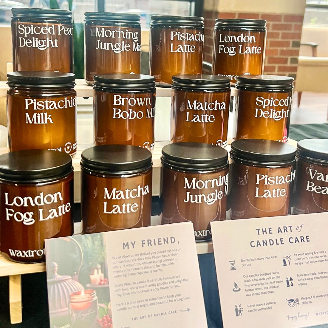 Rows of amber candle jars with scent names like Pistachio Latte and London Fog Latte on a market booth table, with a care card.